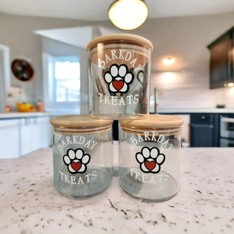 Three glass dog treat jars with paw print logos and bamboo lids on a kitchen counter